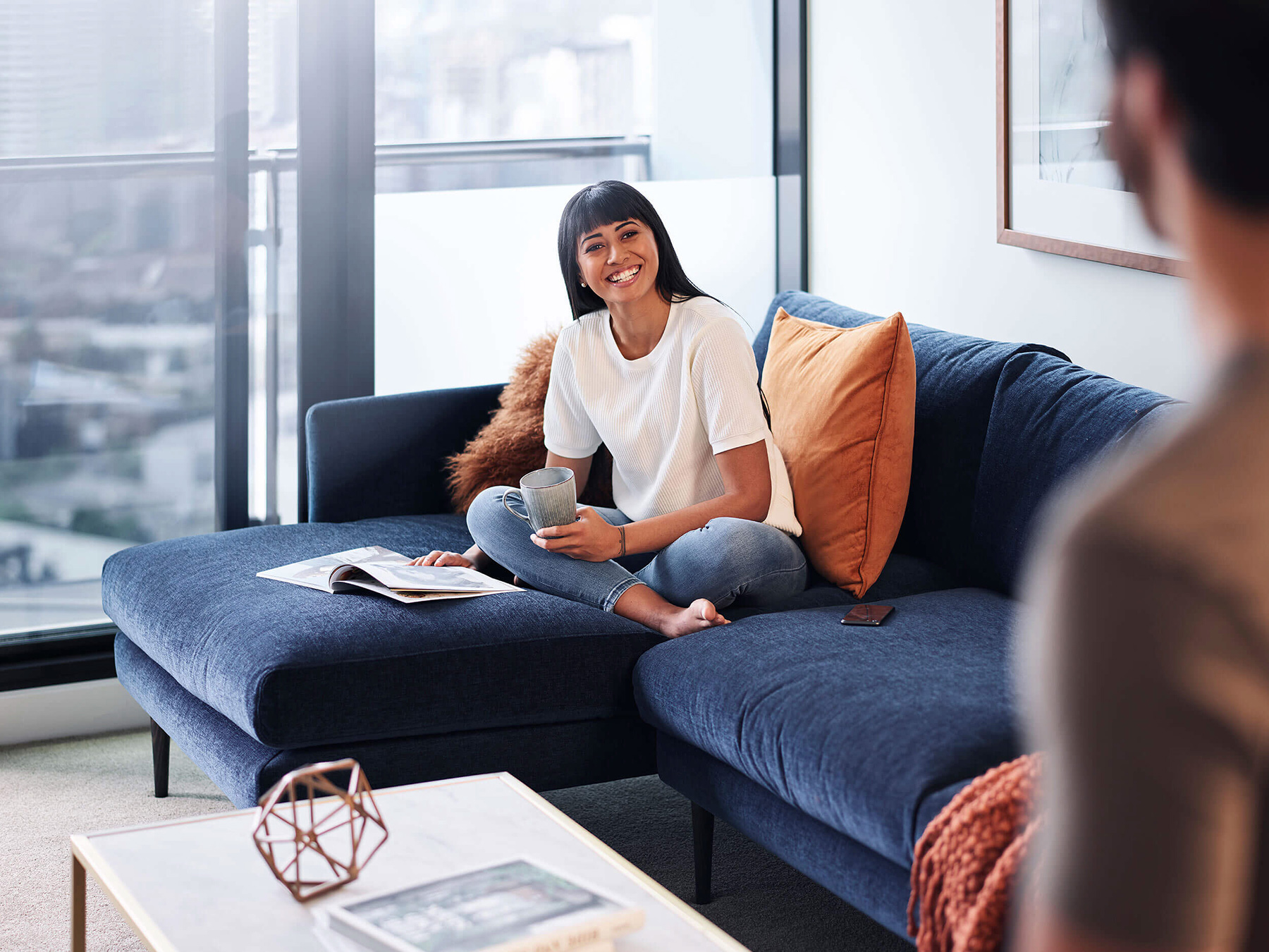 A smiling woman with bangs sits cross-legged on a blue sofa with pillows and a drink, illuminated by natural light from nearby windows.
