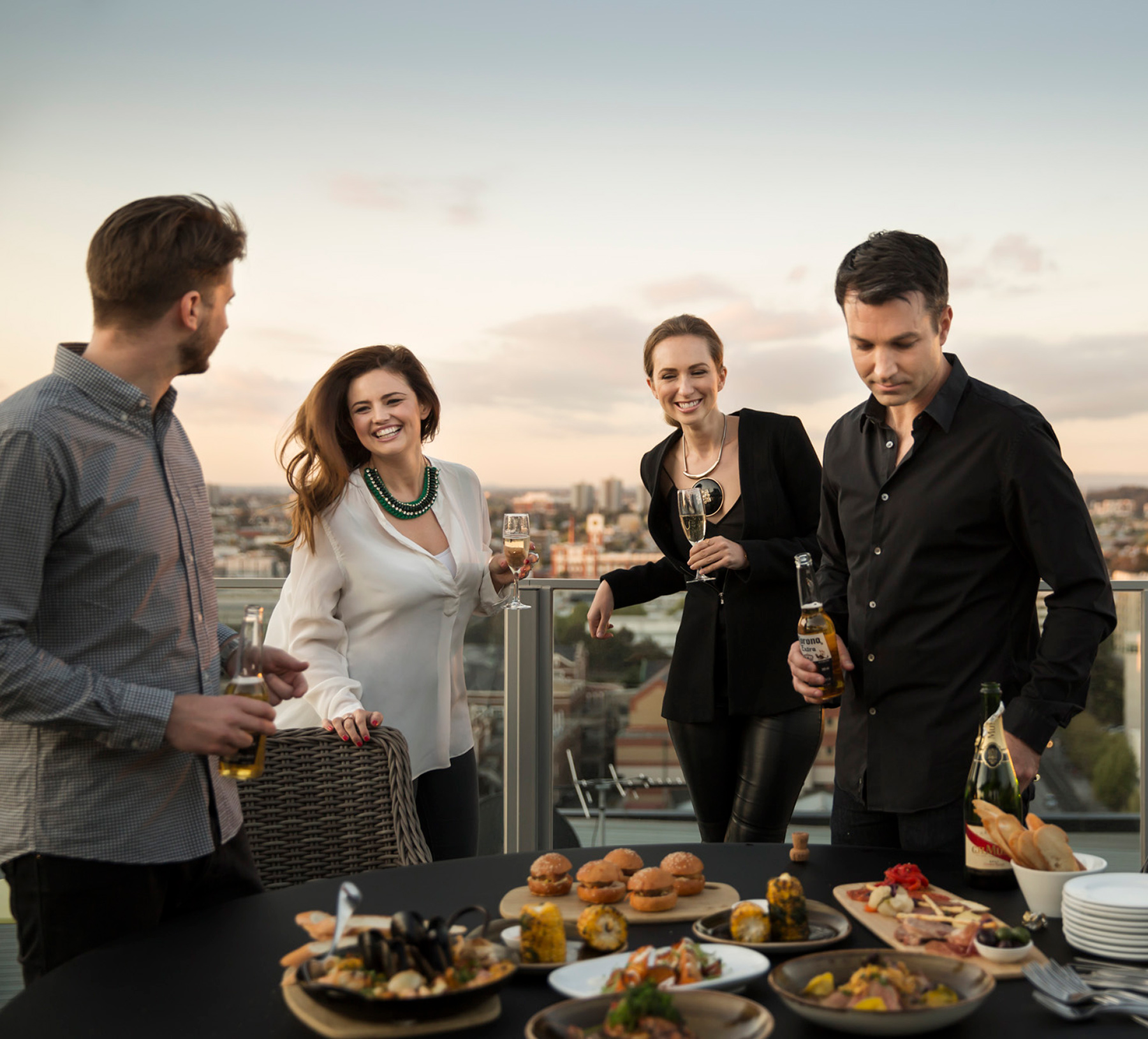 Four people enjoy drinks and appetizers on a rooftop, overlooking a city skyline at dusk, with a table laden with food.