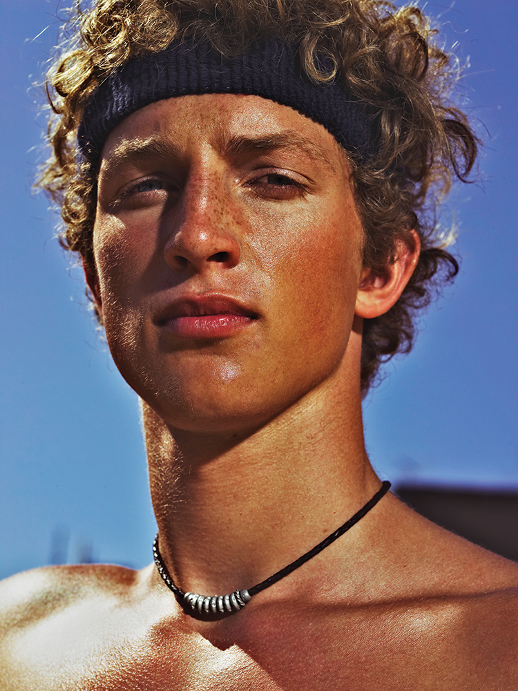 Close-up portrait of a young adult male with curly hair wearing a headband and beaded necklace against a clear blue sky background.