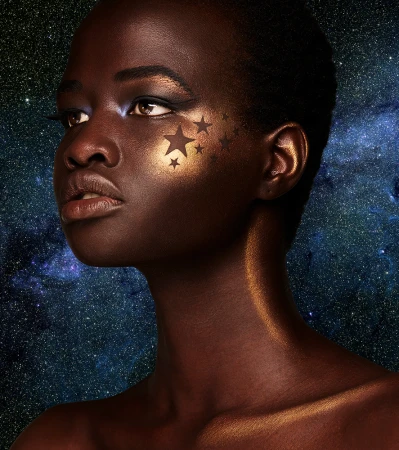 Close-up portrait of a woman with dark skin and dramatic gold makeup and star decals on her face, set against a cosmic background.