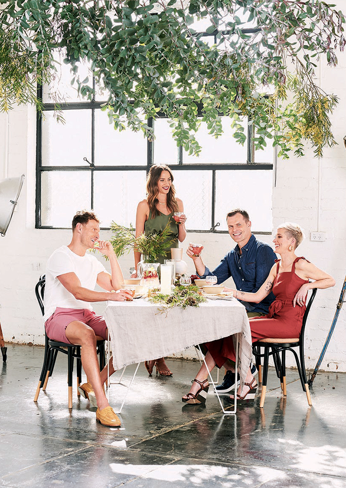 Four people sit at a table with drinks and food, an indoor shot with a large window, greenery, and natural lighting.