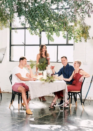 Four people sit at a table with drinks and food, an indoor shot with a large window, greenery, and natural lighting.