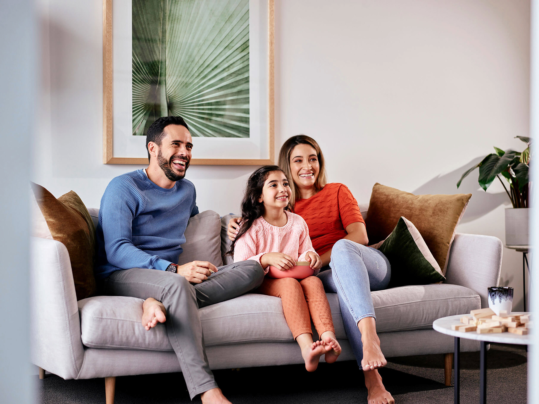 Family of three, including a man, woman, and child, laugh while sitting on a sofa in an indoor living room with neutral tones.