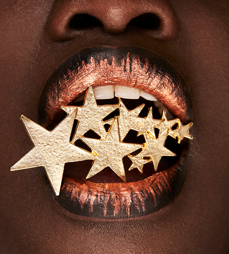 Close-up of a Black woman with bronze-painted lips holding a gold star arrangement in her mouth, showcasing beauty and makeup artistry.