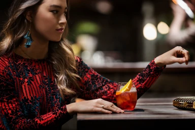 Woman in red and black patterned dress seated at a dark wooden bar with a cocktail featuring an orange slice