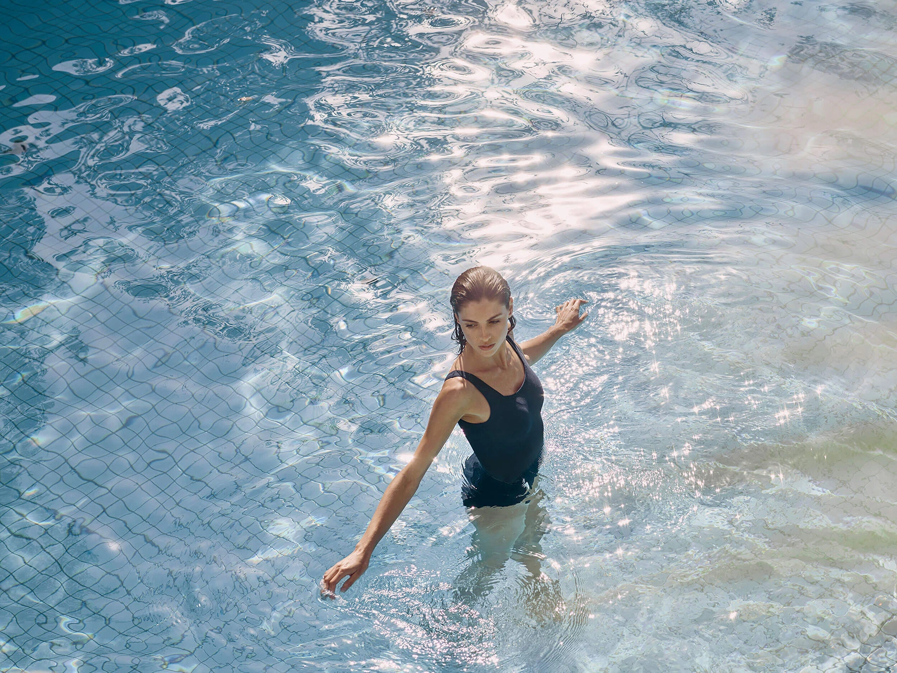 Woman wearing a dark swimsuit standing in a sparkling swimming pool in Melbourne with sunlight reflecting on the water surface.