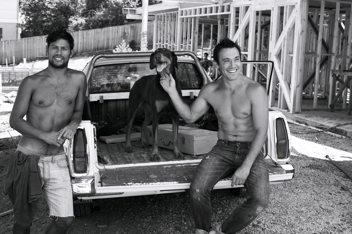 Two shirtless men and a Doberman dog pose by a white pickup truck on a residential construction site in Melbourne.