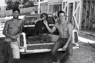 Two shirtless men and a Doberman dog pose by a white pickup truck on a residential construction site in Melbourne.