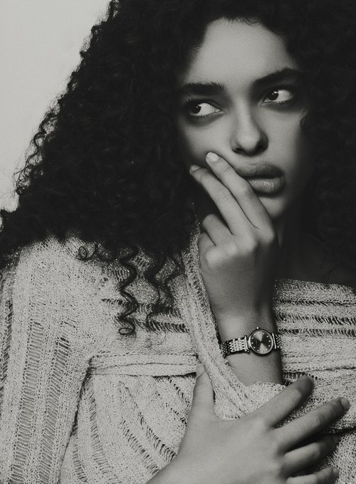 Portrait of an adult woman with curly dark hair, wearing a light-colored open-knit sweater, looking to the side with hand near face.