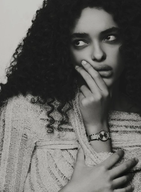 Portrait of an adult woman with curly dark hair, wearing a light-colored open-knit sweater, looking to the side with hand near face.