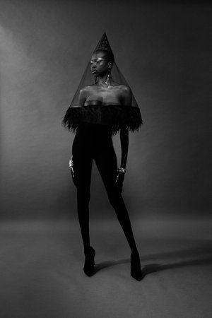 Elegant dark-skinned woman in black fashion ensemble featuring a feathered top, leggings, high heels, and a cone-shaped veil, in studio.