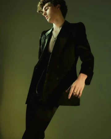 Young man with curly hair wearing a dark velvet suit jacket against a moody gradient background