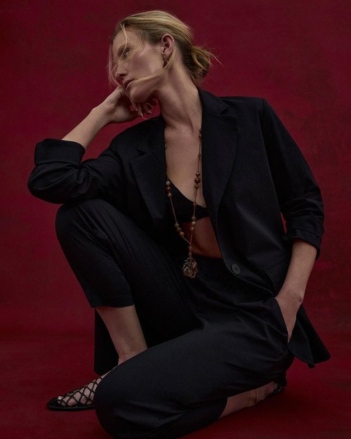 Elegant blonde woman in a black suit and bra, posing in a crouch against a red background in a fashion editorial style portrait.