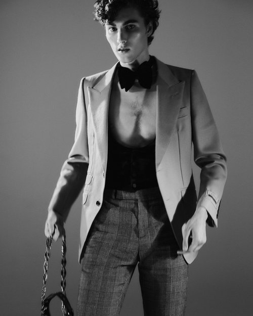 Portrait of a young adult male in a blazer and patterned pants, holding a chain; black and white studio shot, fashion editorial.