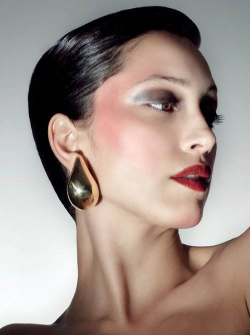Headshot of woman with dark hair and striking makeup wearing a gold teardrop earring in a studio setting with soft, diffused lighting.