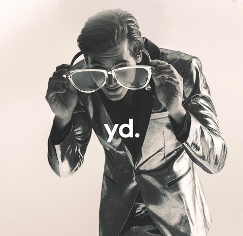 Adult male in a silver suit poses with oversized glasses, staring downward against an off-white background with brand logo, 'yd.' visible.