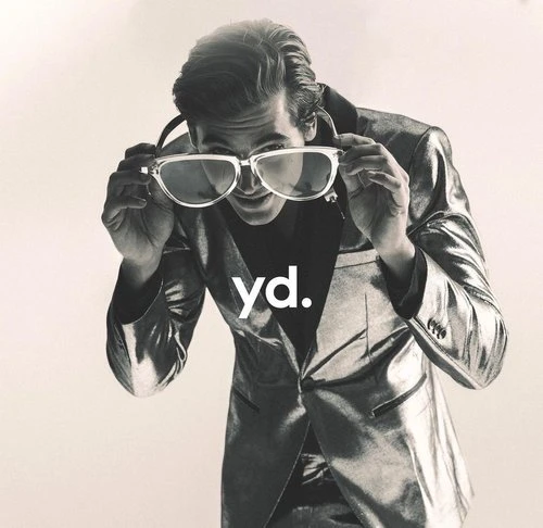 Adult male in a silver suit poses with oversized glasses, staring downward against an off-white background with brand logo, 'yd.' visible.