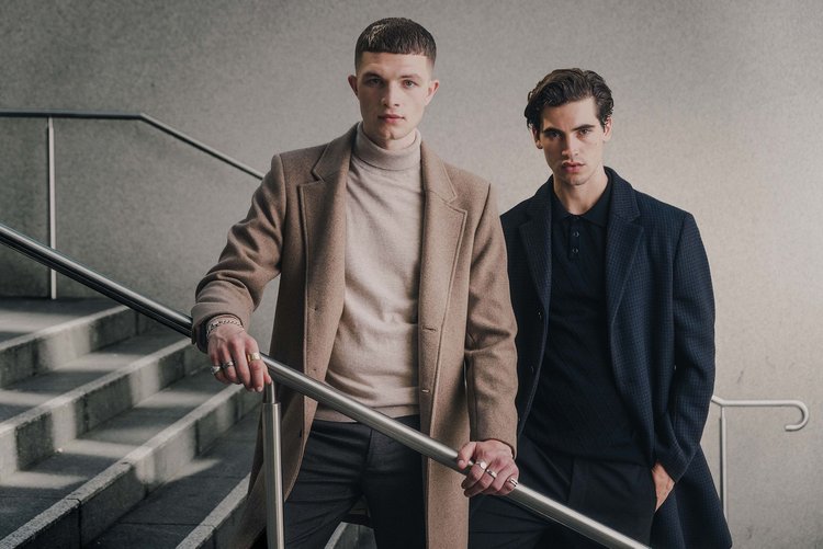Two adult male fashion models in overcoats pose on concrete steps with a metal handrail in an urban setting, demonstrating fall fashion.