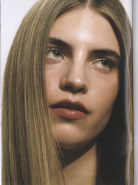 Close-up portrait of a young adult woman with long brown hair, fair skin, and green eyes, looking off-camera against a neutral backdrop.