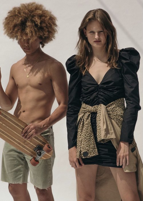 A fashion shot of a male and female model in a studio setting. The male model holds a skateboard and the female model wears a black dress.