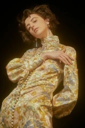 Low angle portrait of a woman wearing a patterned high collar dress against a dark background, hair styled by Darren Summors