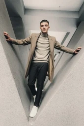A man in a camel coat and turtleneck standing within a triangular architectural structure, styled by hair stylist Darren Summors.