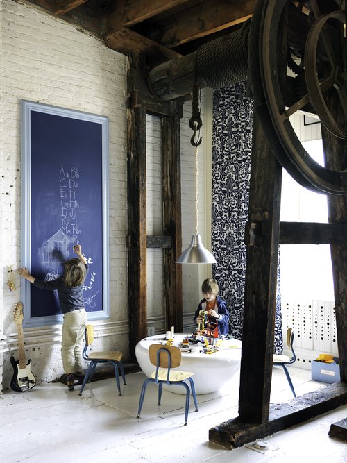 Two young children playing in a brightly lit, industrial-style room with exposed beams, chalkboard, vintage chairs, and modern decor.