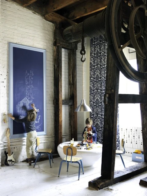 Two young children playing in a brightly lit, industrial-style room with exposed beams, chalkboard, vintage chairs, and modern decor.