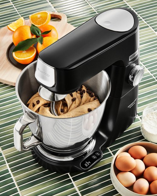 Black stand mixer with dough in bowl and oranges, flour, and eggs on a green tile surface, creating a vibrant cooking scene.