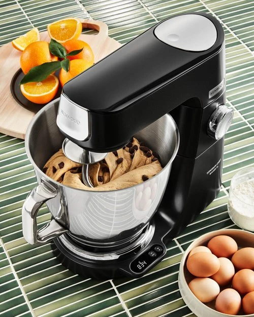 Black stand mixer with dough in bowl and oranges, flour, and eggs on a green tile surface, creating a vibrant cooking scene.