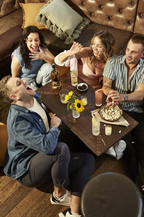 A group of four young adults are gathered around a low table on a wooden floor, laughing and enjoying drinks in a cozy, indoor setting.