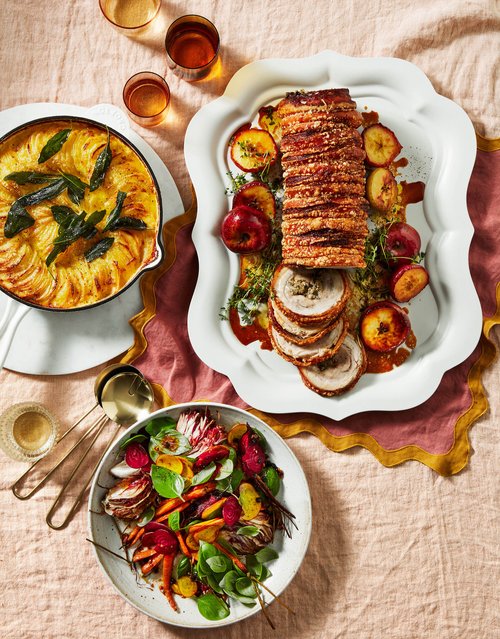 Overhead shot of a porchetta roast, potato gratin, and salad, displayed on a pink linen tablecloth with drinks, suggesting a festive meal.