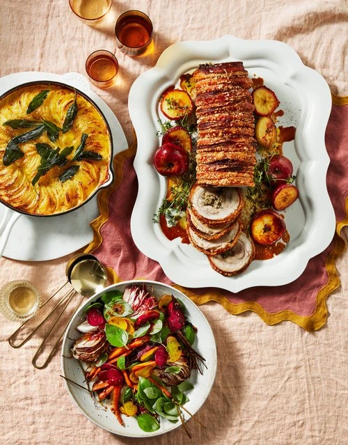 Overhead shot of a porchetta roast, potato gratin, and salad, displayed on a pink linen tablecloth with drinks, suggesting a festive meal.