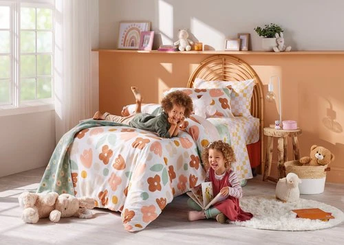 Two young children, a boy and a girl, are in a brightly lit room on and around a bed, one lying and one reading a book.