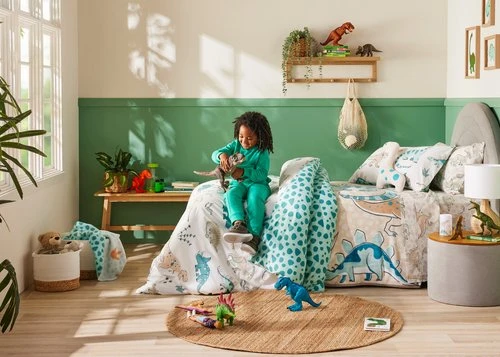 Child in green pajamas sits on a dinosaur-themed bed in a brightly lit room, playing with a toy dinosaur. Dinosaur toys are also on the floor.