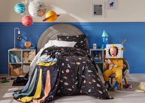 Toddler in astronaut costume plays in bedroom decorated with space-themed bedding and decor against a blue wall with framed pictures.