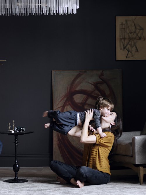 Woman playfully lifts a toddler indoors in a cozy home, showcasing dark walls, carpet flooring, modern lighting, and home decor.