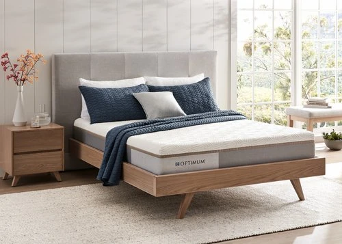 Bedroom interior featuring a bed with a white mattress, blue pillows and blanket, a wooden headboard, and a bedside table with flowers.