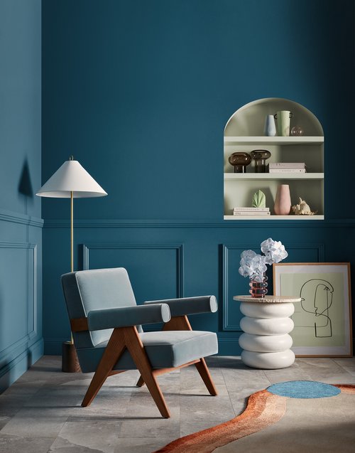 Interior design featuring a light blue mid-century modern armchair, a white side table with flowers, and a framed drawing in a blue room.