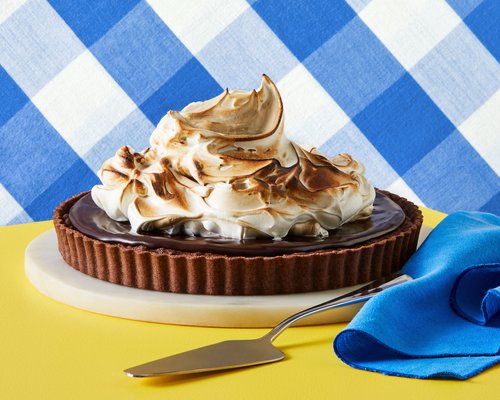 Chocolate tart topped with toasted meringue on a marble and yellow surface, set against a blue and white checkered backdrop.
