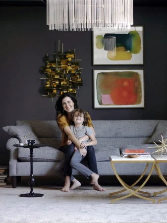 Mother and child sit barefoot on a grey sofa in a home interior with abstract art, modern lighting, and contemporary decor.