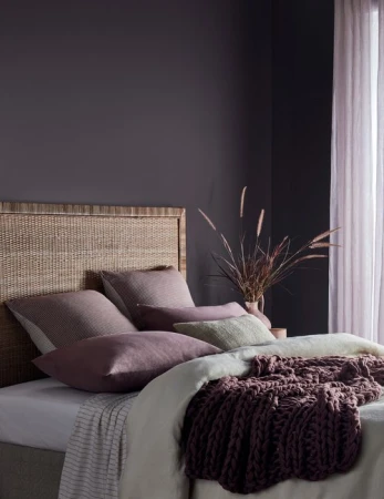 Cozy bedroom interior with a woven rattan headboard, layered pillows in muted tones, and a dark plum wall.