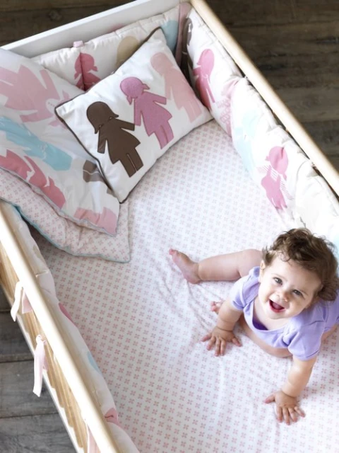 Infant with a cheerful expression crawls in a wooden crib with patterned bedding and pillows. Crib features safety bumper pads.