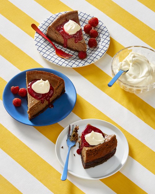 Three slices of chocolate cheesecake with raspberries, red sauce, and whipped cream on colorful plates, set on a yellow striped tablecloth.