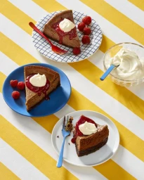 Three slices of chocolate cheesecake on colorful plates with whipped cream and fresh raspberries on a yellow striped background.