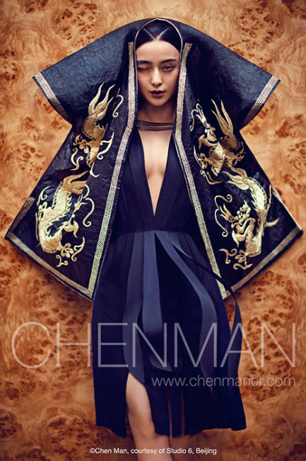 An Asian woman posing dramatically in a deep V-neck dress with an embroidered cape, captured in a fashion-style shot against a wood-grain backdrop.