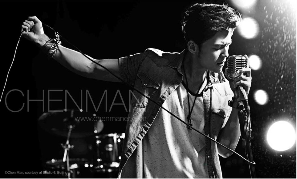 Black and white portrait of a male singer performing live with a vintage microphone in front of stage lighting and a drum set.