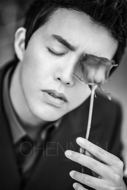 Monochrome portrait of an adult male with closed eyes holding a rose, wearing a suit and tie, with soft lighting.