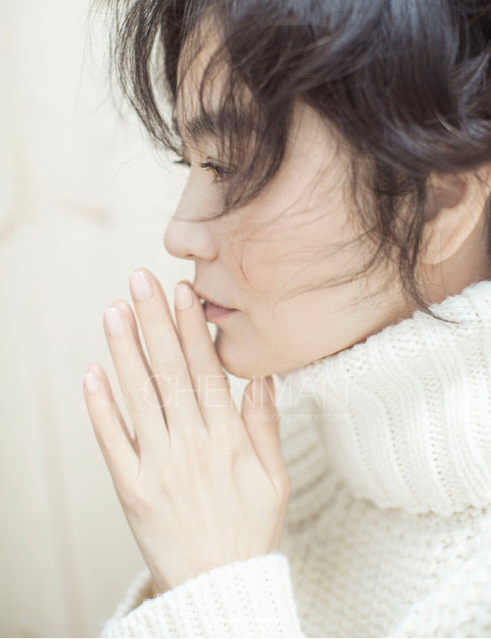 Portrait of a woman with dark hair in a white turtleneck sweater, her hands near her face, soft lighting, and a neutral background.