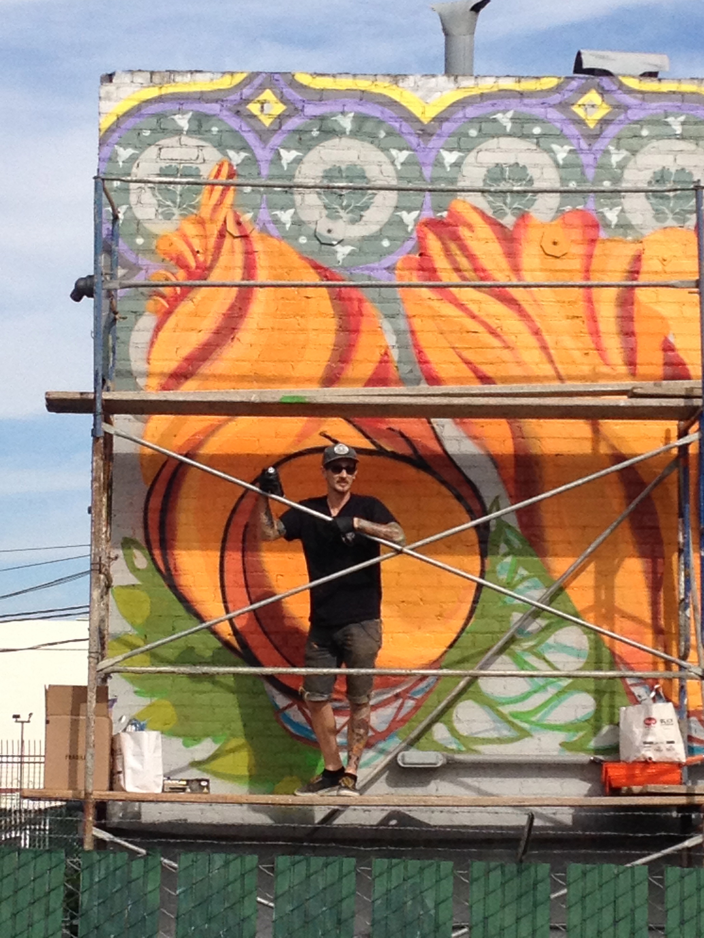 Artist stands on scaffolding in front of a large orange and green floral mural on a brick building, holding a spray can.